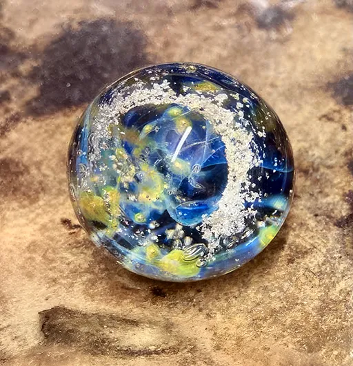 Eine blau-gelbe Glasmurmel mit wirbelnden Mustern auf brauner Oberfläche.
