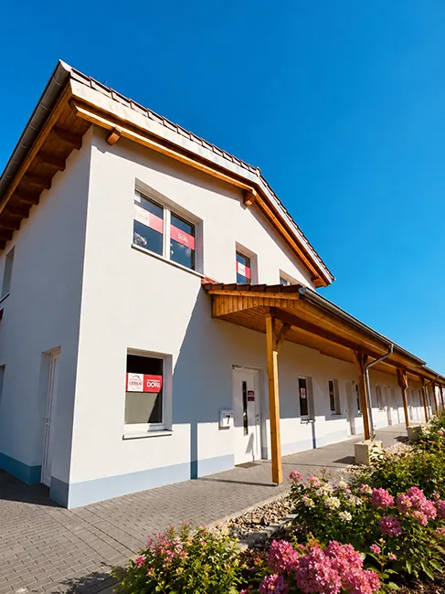 Ein weißes Gebäude mit Holzdach, davor Blumen und darüber ein strahlend blauer Himmel.